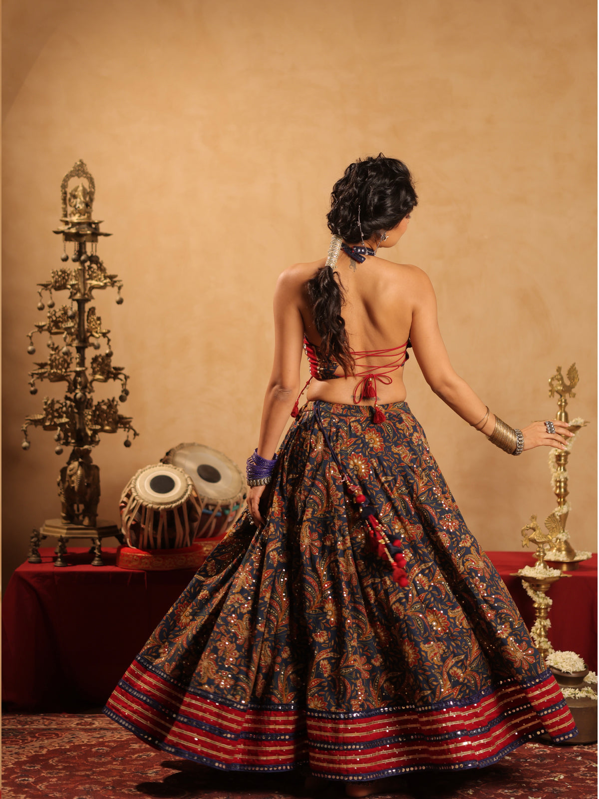 Rear full-length standing view of a model in the Rangrez Red & Blue Heritage cotton chaniyacholi set showing the expansive patterned flare, metallic striped borders, backless tie-up blouse, and long braided hair beside traditional brass lamps.