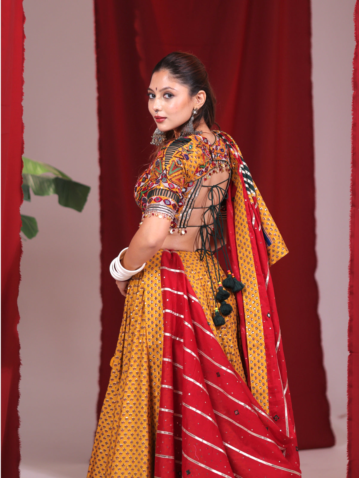 Rear side-angle view of a model wearing a mustard yellow printed cotton chaniya choli set highlighting the open-back tie-up dori blouse and draped red striped dupatta for Navratri garba.