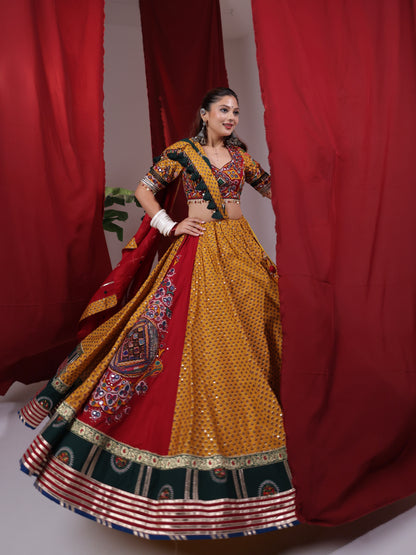 Front three-quarter view of a model in a mustard yellow printed cotton chaniya choli set holding out the flared ghaghra to highlight the dark green arch border and crimson red panel for Navratri garba.