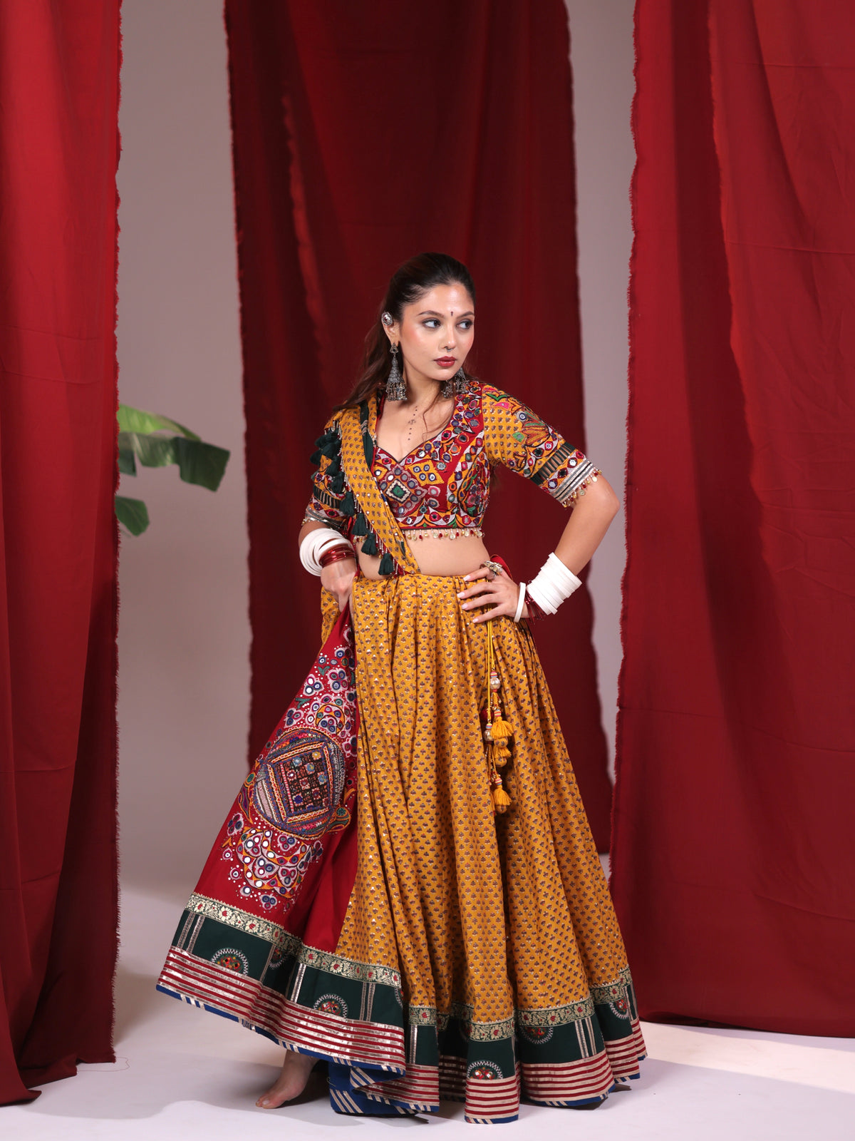 Front standing view of a model in a mustard yellow printed cotton chaniya choli set highlighting the draped crimson red dupatta with mirror-work patch, green hem borders, and waist tassels for Navratri garba.