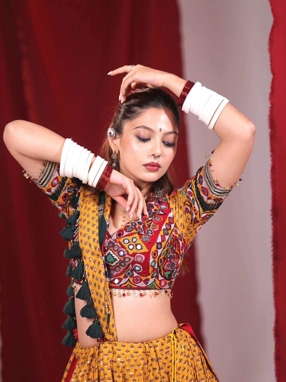 Front half-portrait of a model wearing a mustard yellow cotton chaniya choli set highlighting the multicolor embroidered choli, hanging dark green tassel latkans, and traditional white bangles for Navratri garba.