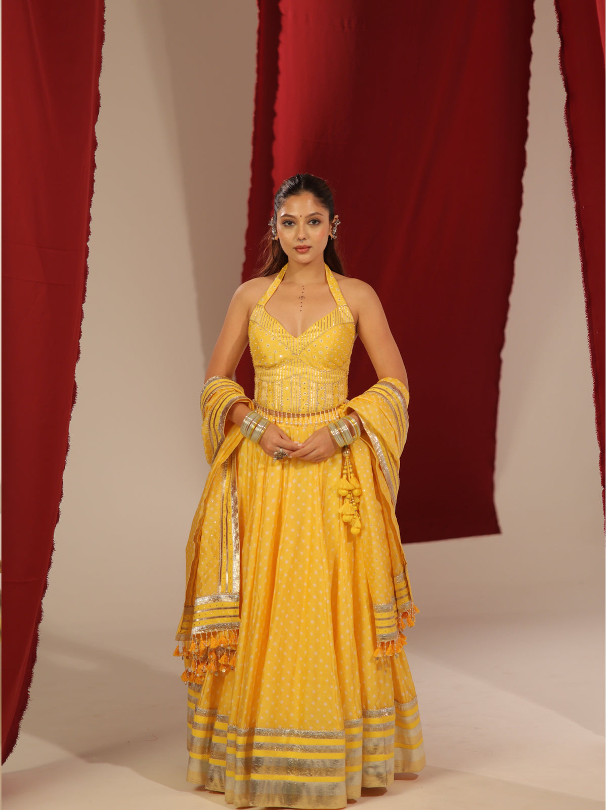 Front full-length view of a model wearing the Peeli Haldi yellow printed halter chaniyacholi set featuring a matching draped dupatta with tassels and silver gota border against red curtains for Navratri garba.