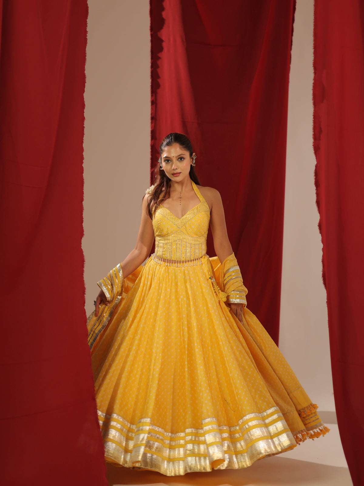 Front full-length view of a model wearing the Peeli Haldi vibrant yellow halter chaniyacholi set featuring a voluminous flared skirt with broad silver gota borders and embroidered halter bodice against red curtains for Navratri garba.