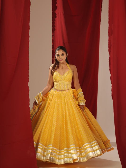 Front full-length view of a model wearing the Peeli Haldi vibrant yellow halter chaniyacholi set featuring a voluminous flared skirt with broad silver gota borders and embroidered halter bodice against red curtains for Navratri garba.